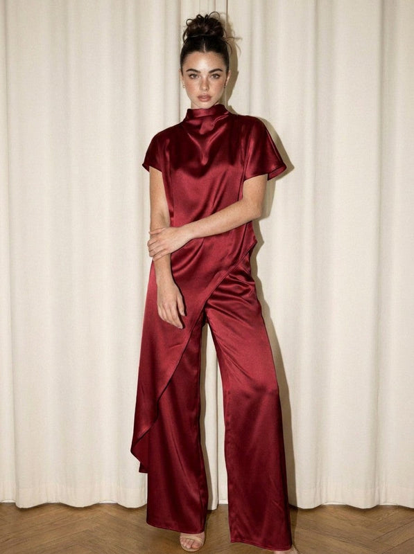 Model seated on stool wearing champagne version of satin cape-back mock neck jumpsuit